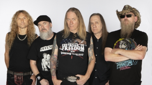 Five male rock band members pose together in black shirts, long hair, and tattoos against a white background.