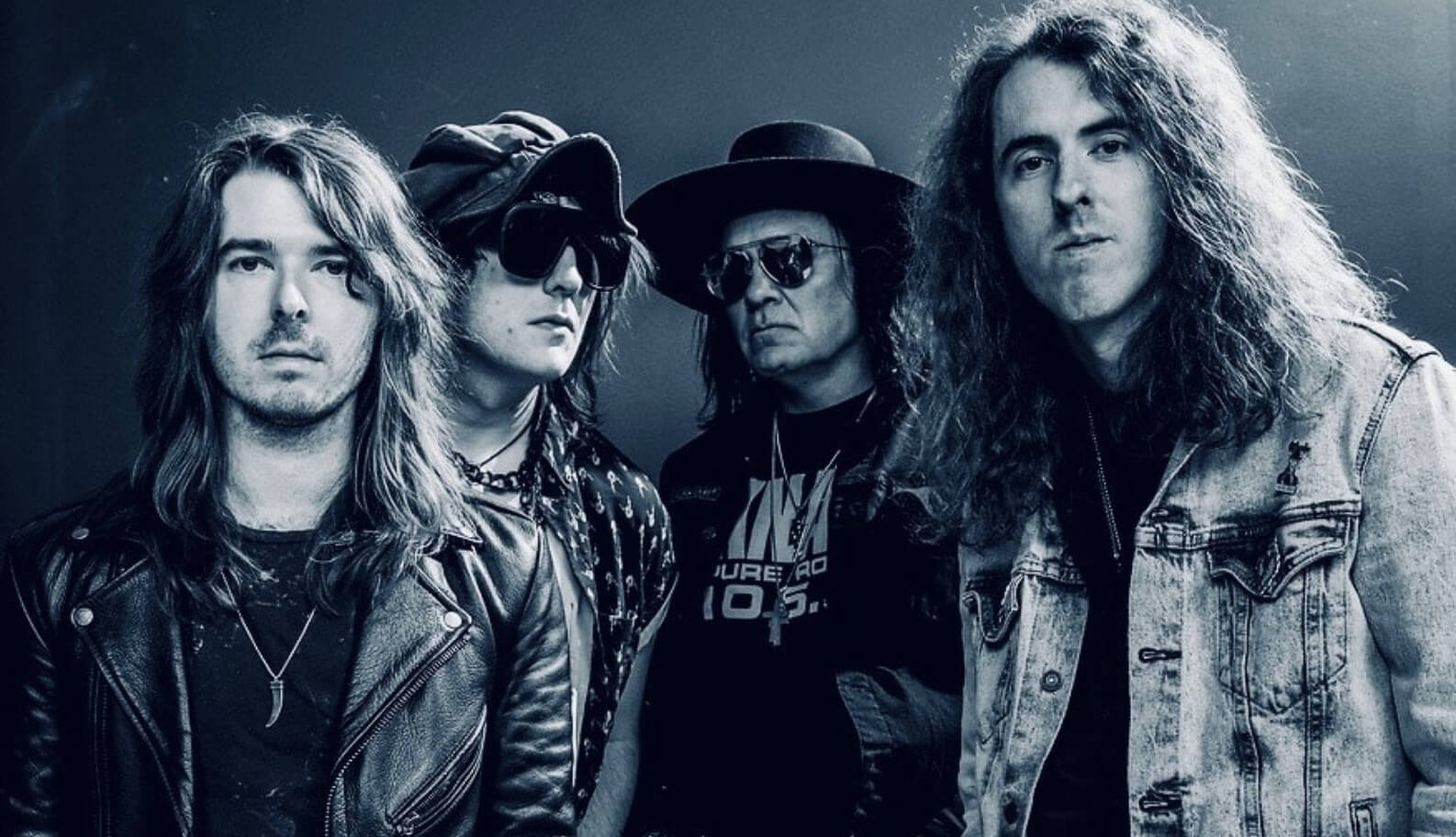 | The Mountain Music Festival Four male rock band members pose together in a blue-toned studio portrait wearing leather and denim jackets.