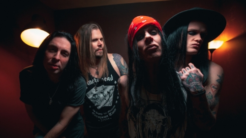 Four band members posing in a moody, red-lit room; three men with long hair, one wearing a red bandana, all wearing dark shirts with visible tattoos, conveying a rock/metal band vibe.