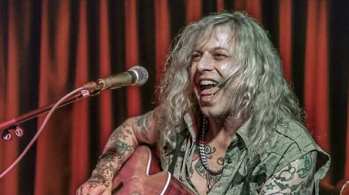 Male guitarist with long, wavy hair and visible tattoos, playing an acoustic guitar and singing into a microphone on stage with red curtains behind him