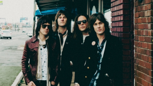 Four men with long hair and sunglasses posing on a city sidewalk outside a brick building.