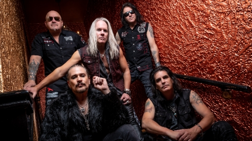 Five male rock band members pose on a stairwell against a red, textured wall, wearing black vests and leather.