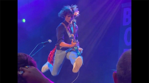 Guitarist mid-air jump on a dimly lit stage with blue/purple lights, playing an electric guitar; audience hands visible in the foreground.